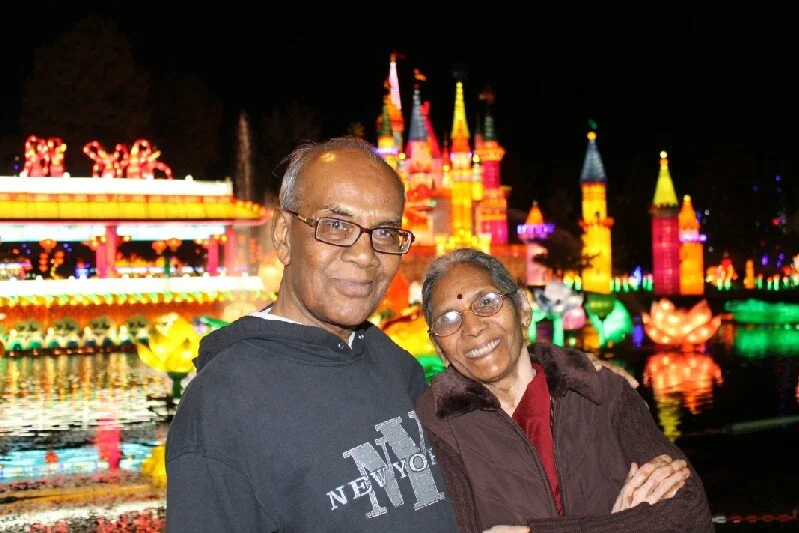 A smiling older man and woman pose in front of a brightly lit, colorful castle display at night, with reflections on the water.