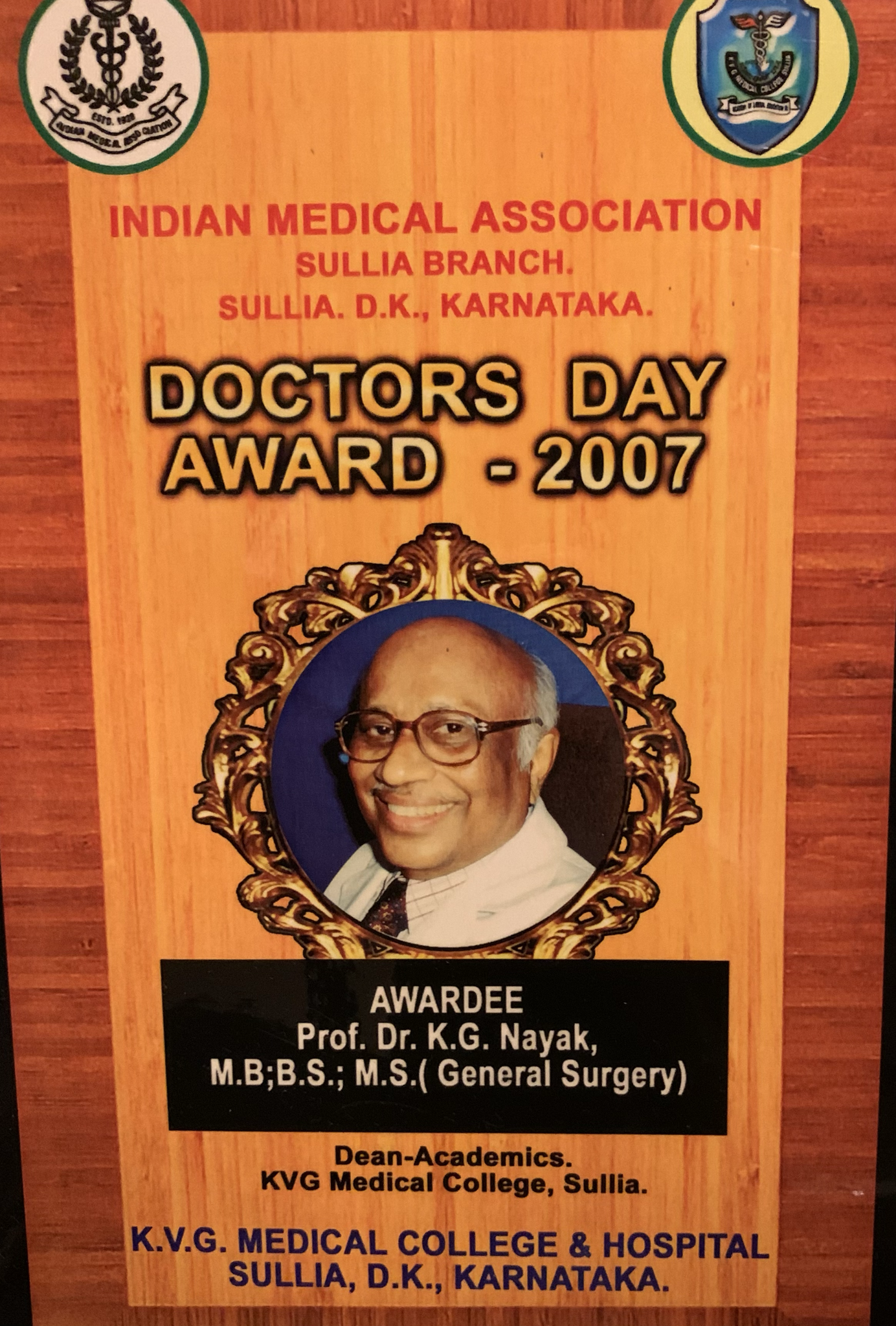 A award plaque from the Indian Medical Association Sullia branch in Karnataka. It features a photo of an elderly man with glasses, smiling, in a gold ornate frame. The plaque details the award given in 2007 to Professor Dr. K.G. Nayak, a senior general surgeon. The background is wood-textured.