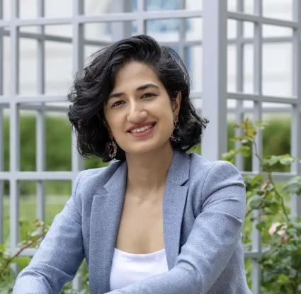 A woman with dark curly hair smiling, wearing a light blue blazer and white top, standing in a greenhouse with green plants and a metal barred structure in the background.