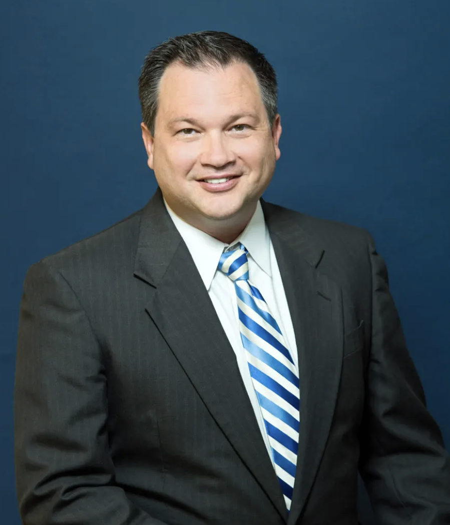A smiling man wearing a dark suit, white shirt, and striped blue and white tie poses against a blue background.