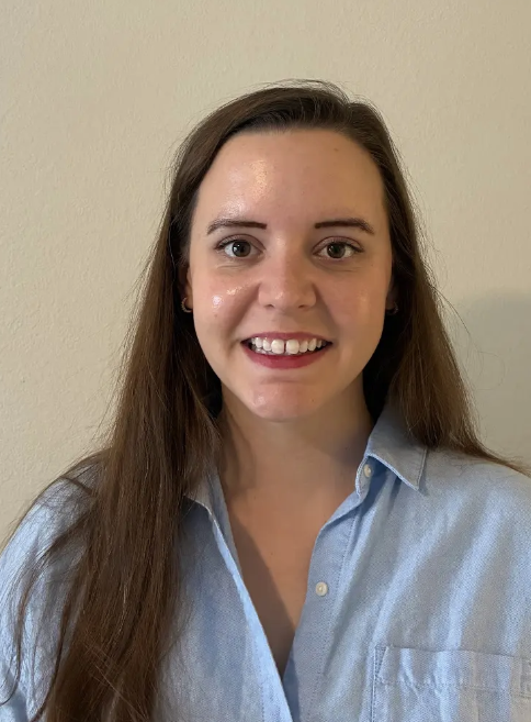 Portrait of a young woman with long brown hair, smiling, wearing a light blue button-down shirt against a plain beige wall.