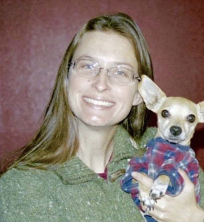 Woman smiling, holding a small dog wearing a plaid shirt, against a red background.