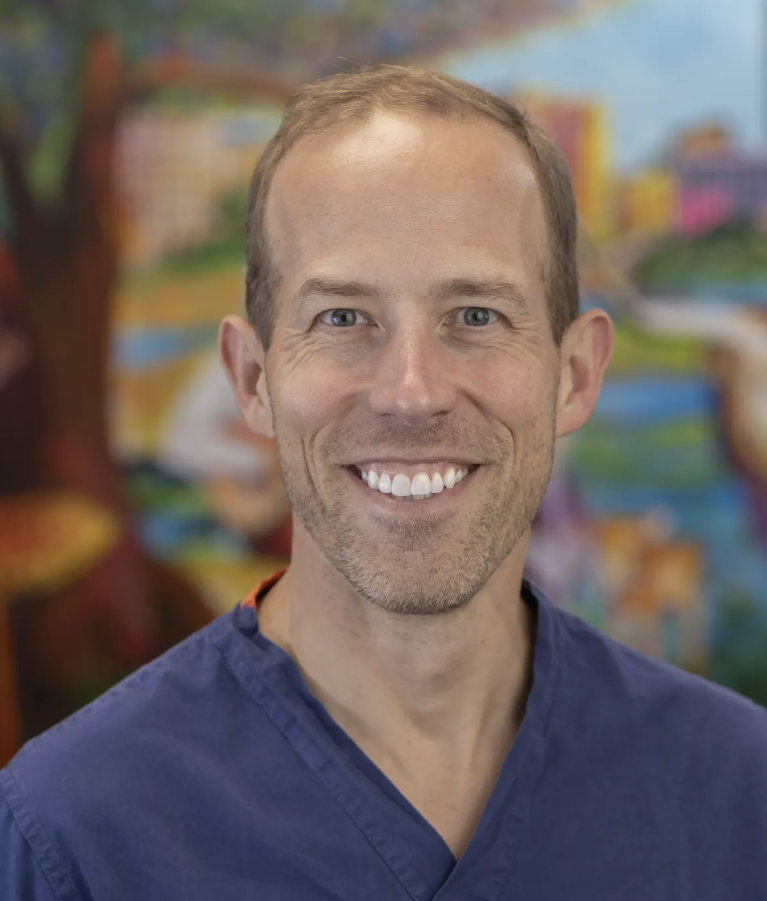 Smiling man with short light brown hair wearing a navy blue shirt, standing in front of a colorful, out-of-focus background.
