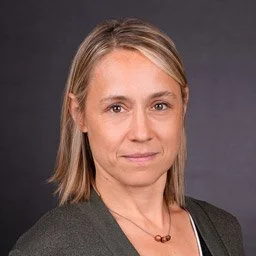 A woman with blonde hair and a neutral expression, wearing a dark blazer and necklace, posed against a dark background.
