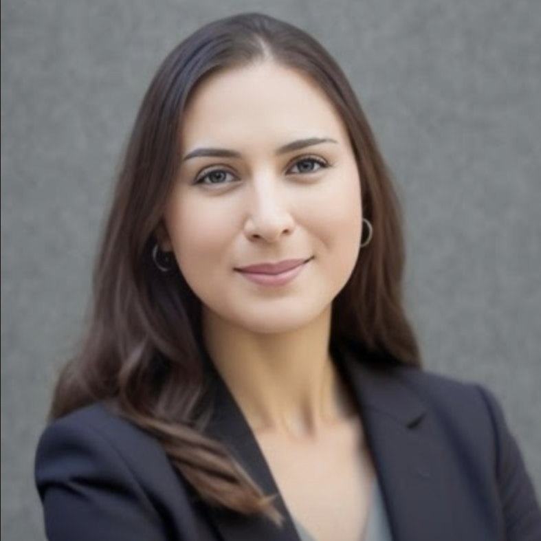 A woman with long brown hair, wearing a black blazer, smiling at the camera, against a gray background.