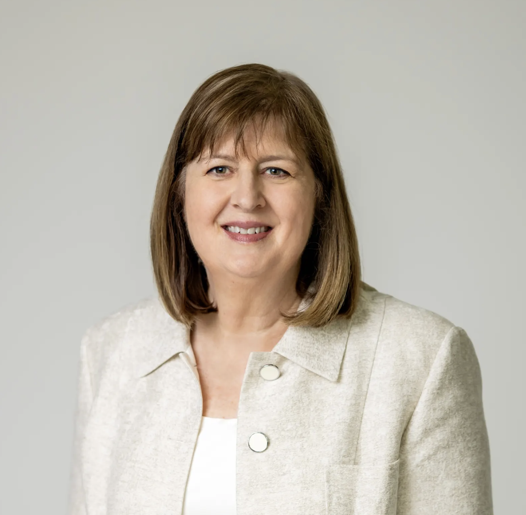 Portrait of a middle-aged woman with brown hair, smiling, wearing a cream-colored blazer and white top, against a plain light background.