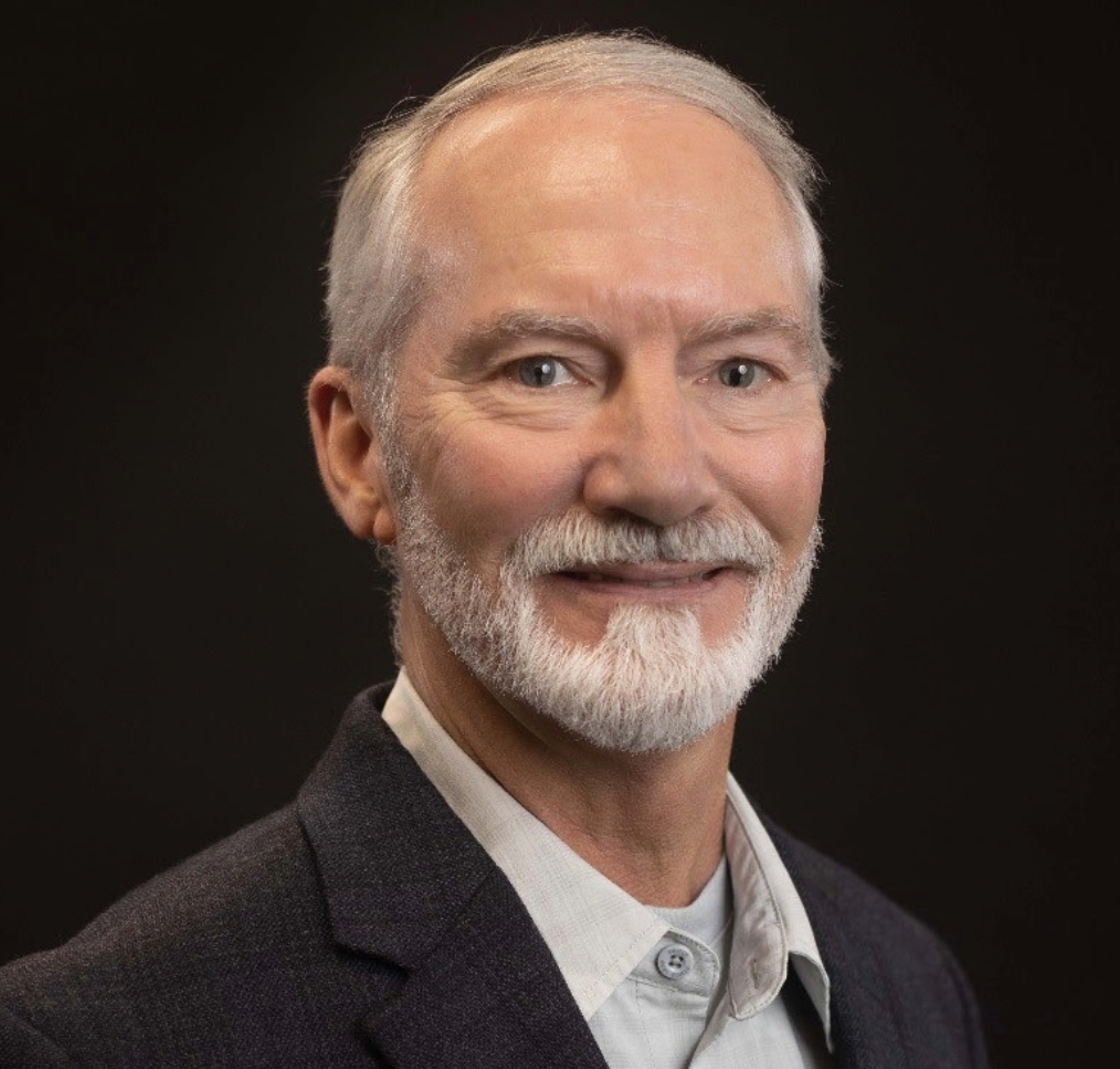 Headshot of an older man with gray hair, beard, and mustache, wearing a dark blazer and light-colored shirt against a dark background.