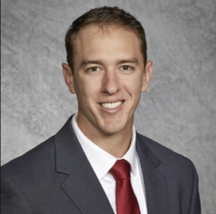 Portrait of a young man wearing a dark suit, white shirt, and red tie, smiling against a gray textured background.
