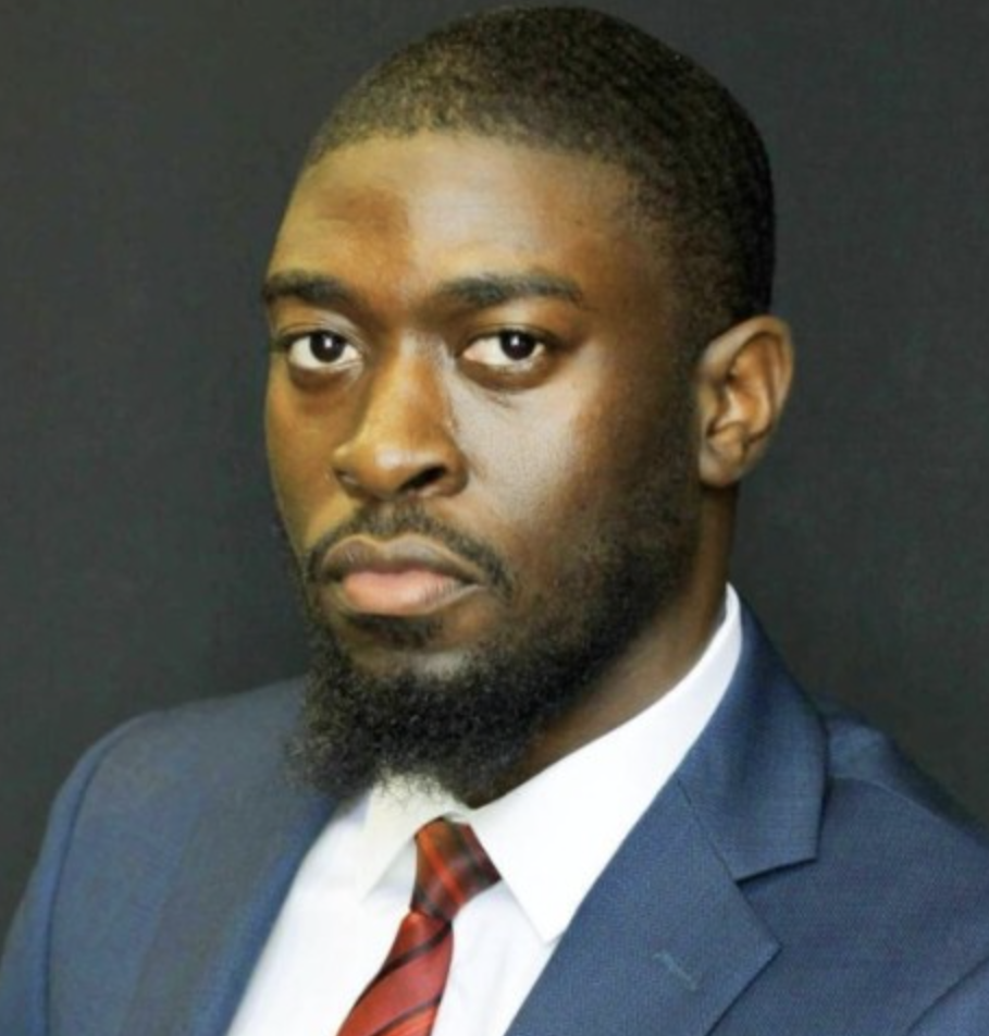 A young man with dark skin, short black hair, and a beard, wearing a blue suit, white shirt, and red striped tie against a black background.