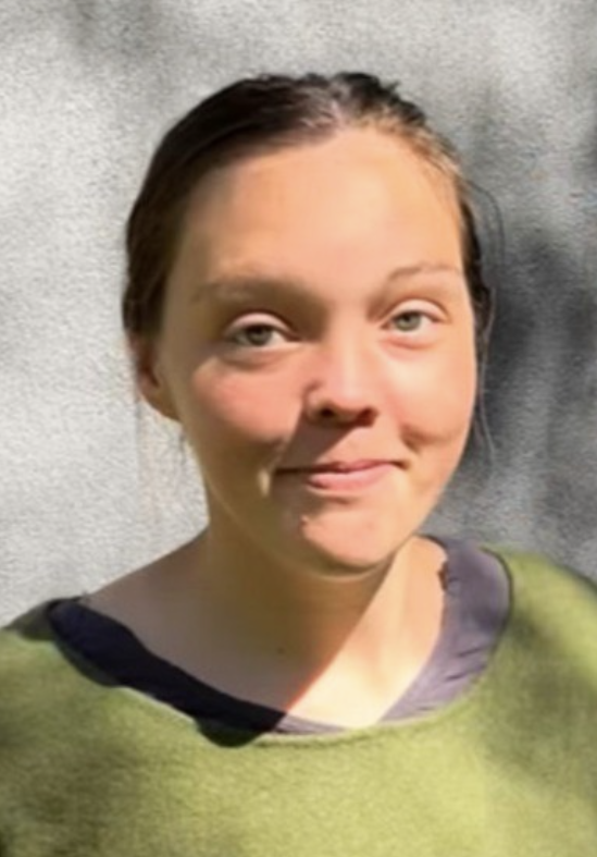 A woman with light skin and brown hair pulled back, smiling softly, standing outdoors against a gray wall, wearing a green top with a dark collar.