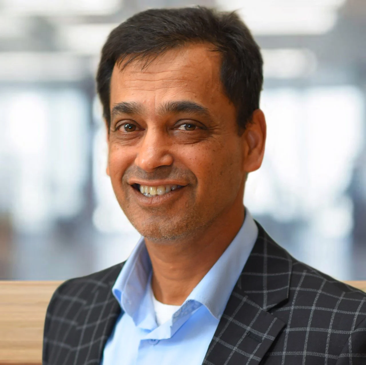 Close-up of a smiling middle-aged man with short black hair, wearing a light blue shirt and a dark checkered blazer, in an indoor setting with blurred background.