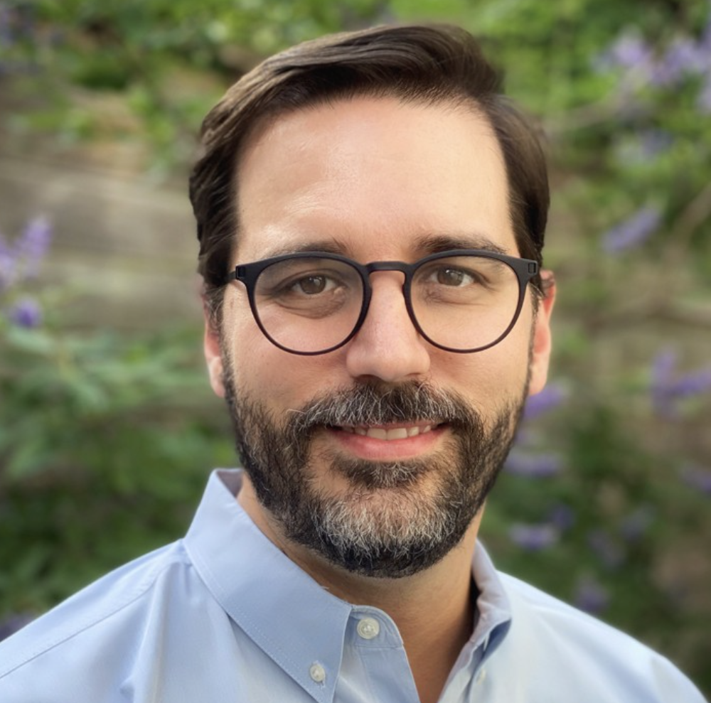 A man with dark hair, glasses, and a beard smiling outdoors with green and purple foliage in the background.