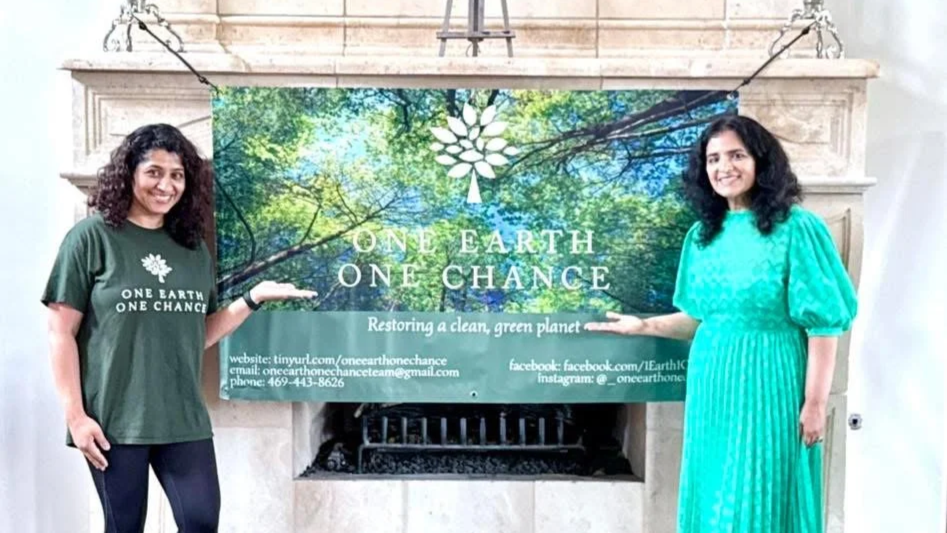 Two women standing in front of a banner that reads "ONE EARTH ONE CHANCE" and features a forest scene. One woman is wearing a dark green T-shirt with the same logo and text, and the other is wearing a bright turquoise dress. They are smiling and pointing to the banner, which promotes restoring a clean, green planet.
