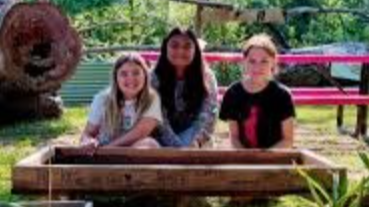 Three young females sitting at a wooden table outdoors, with a large plush bear to the side and trees in the background.