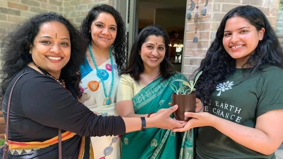 Four women smiling and holding a potted plant together outdoors, standing in front of a brick building.