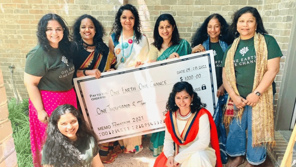 Group of women, with one woman seated in front wearing traditional Indian attire, holding a large ceremonial check addressed to "One Earth One Chance" for $1,000, surrounded by other women standing, some wearing t-shirts with the organization's slogan, in an outdoor setting.