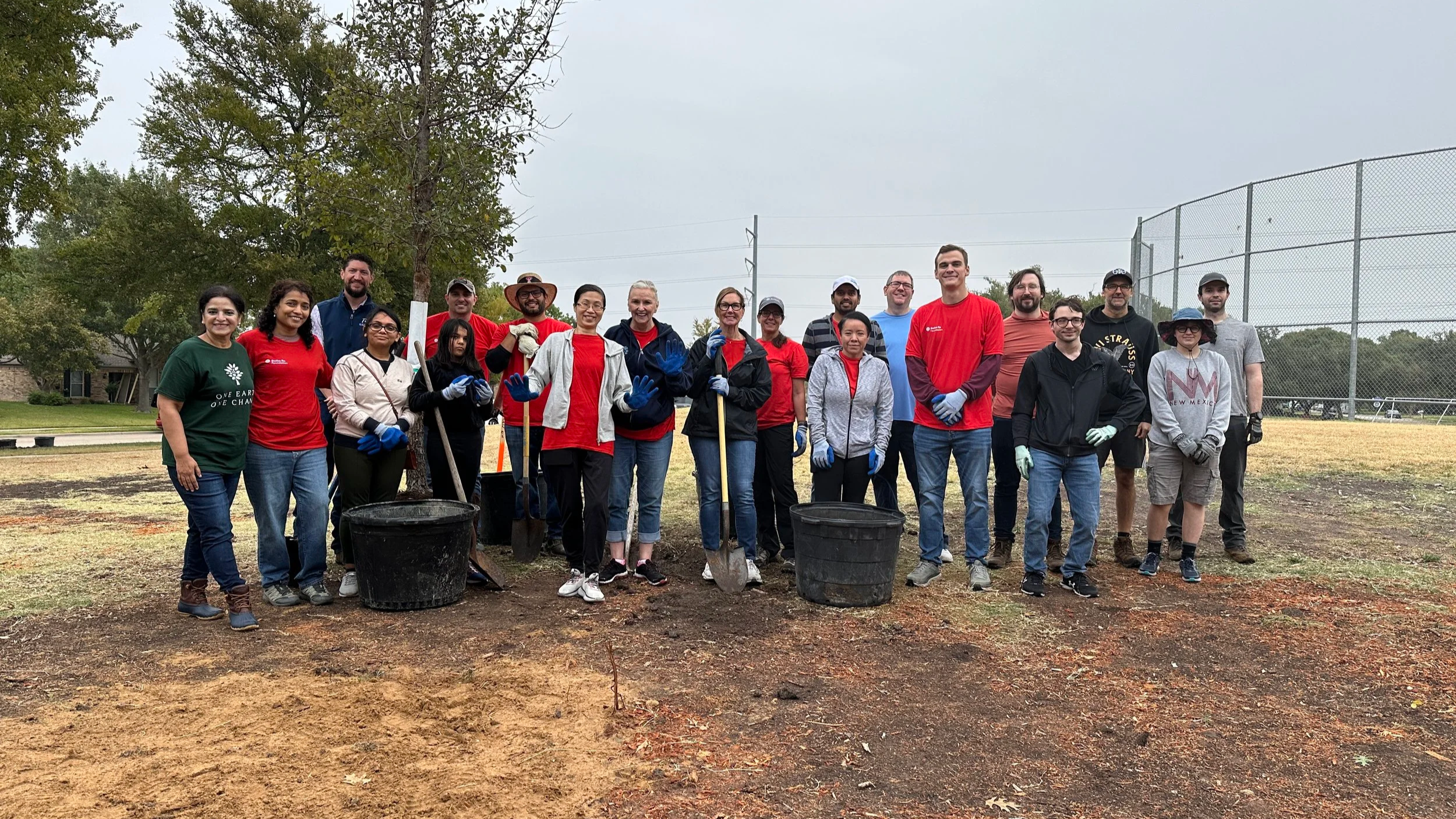 Group of people participating in a community tree planting event, holding tools and posing outdoors near a baseball field and trees.