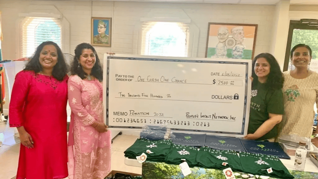 Five women standing behind a large mock check donation in a room with artwork and a table with green and navy T-shirts.
