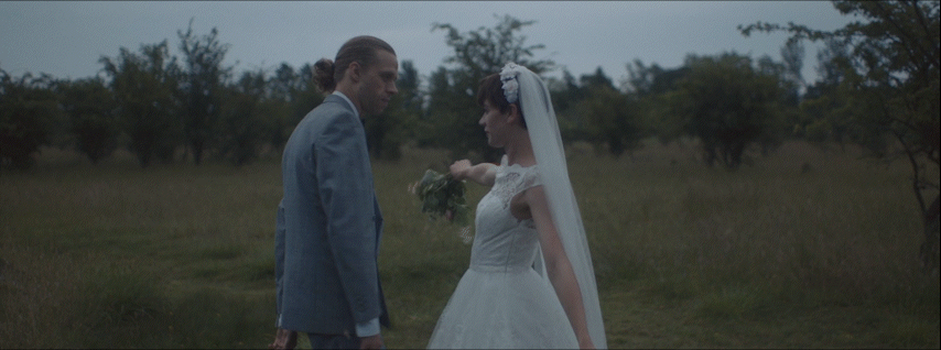 A bride in a white wedding dress and veil holding a bouquet facing a groom in a gray suit in a grassy field with trees in the background.