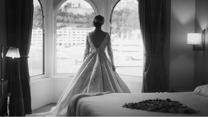A woman in a wedding dress standing by a large bay window, looking outside in a hotel room.