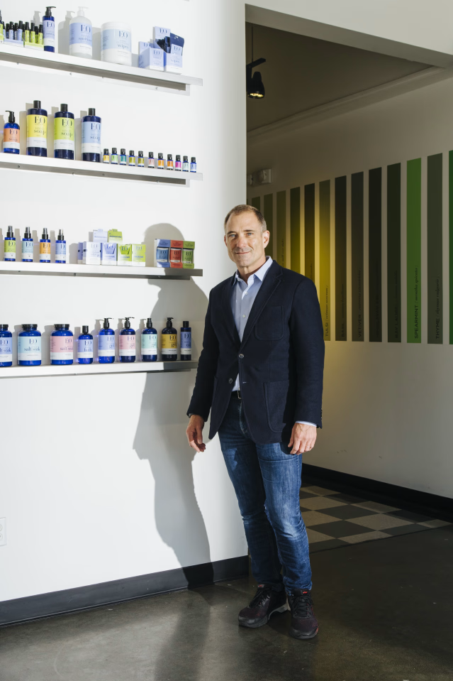 A man named Tom Feegel stands next to shelves displaying various health and wellness products in a retail store or clinic.