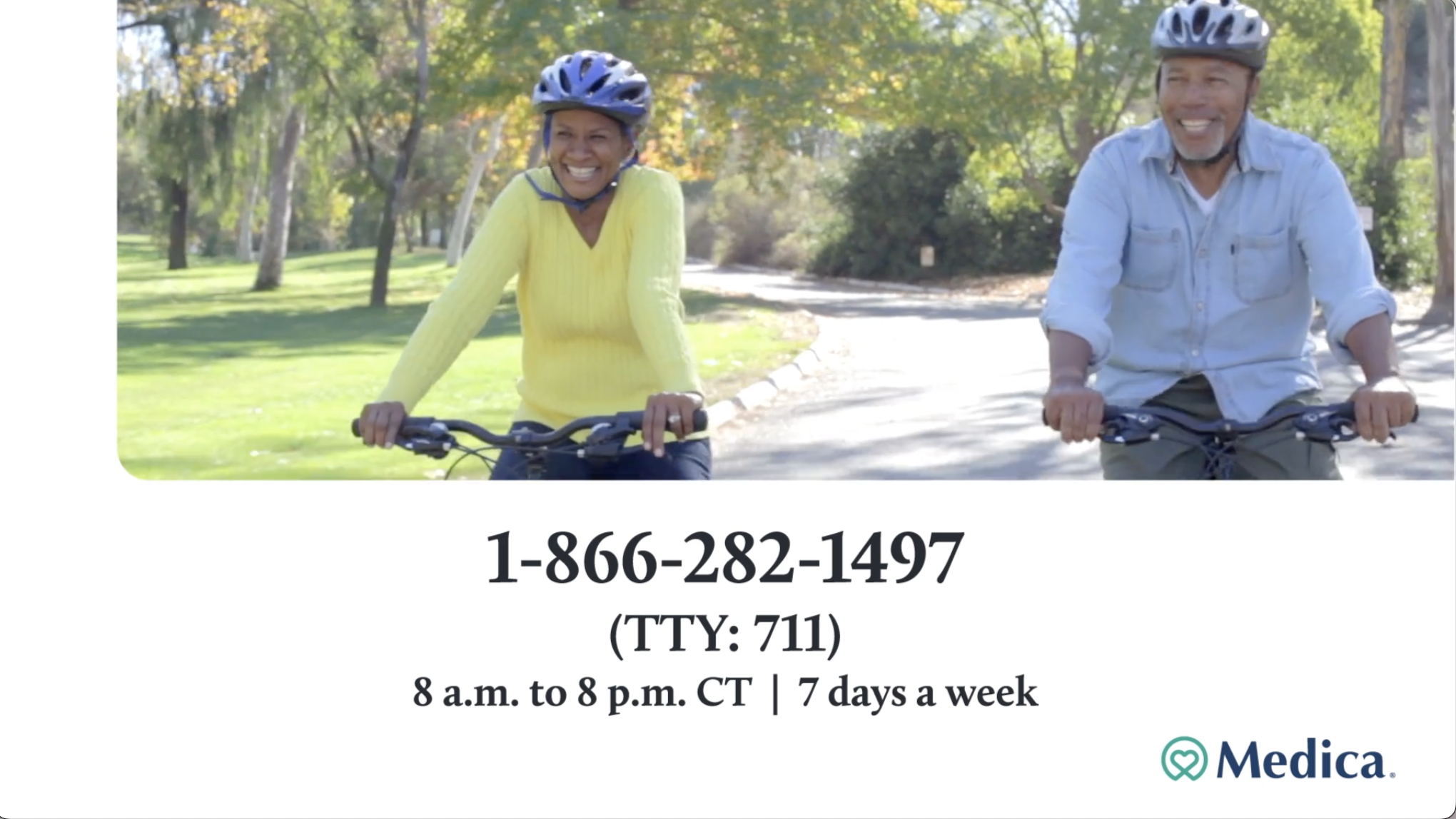 Two smiling people riding bicycles in a park with green trees, wearing helmets, one wearing a yellow sweater and the other wearing a light blue shirt.