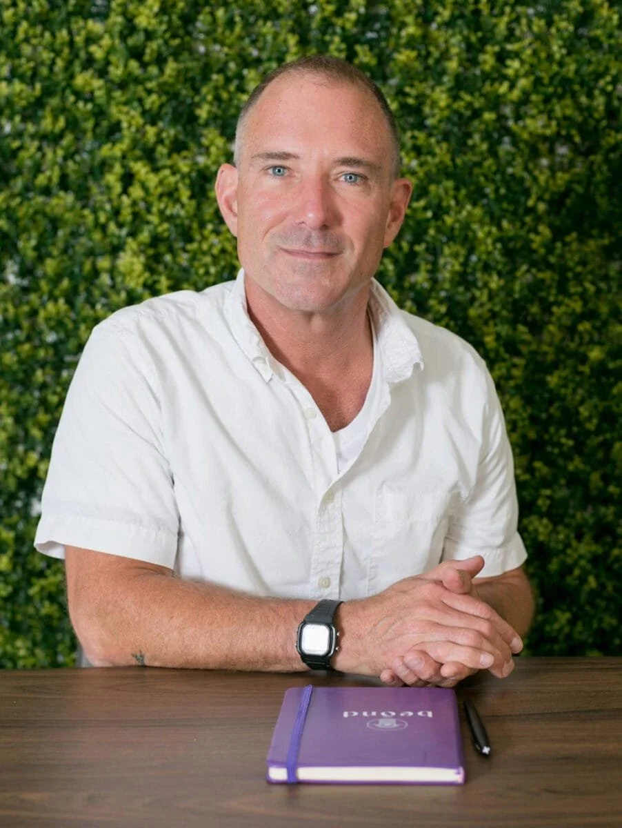 A man named Tom Feegel with short hair and a slight smile, wearing a white short-sleeved button-up shirt, is sitting at a wooden table with a purple notebook and a black pen.