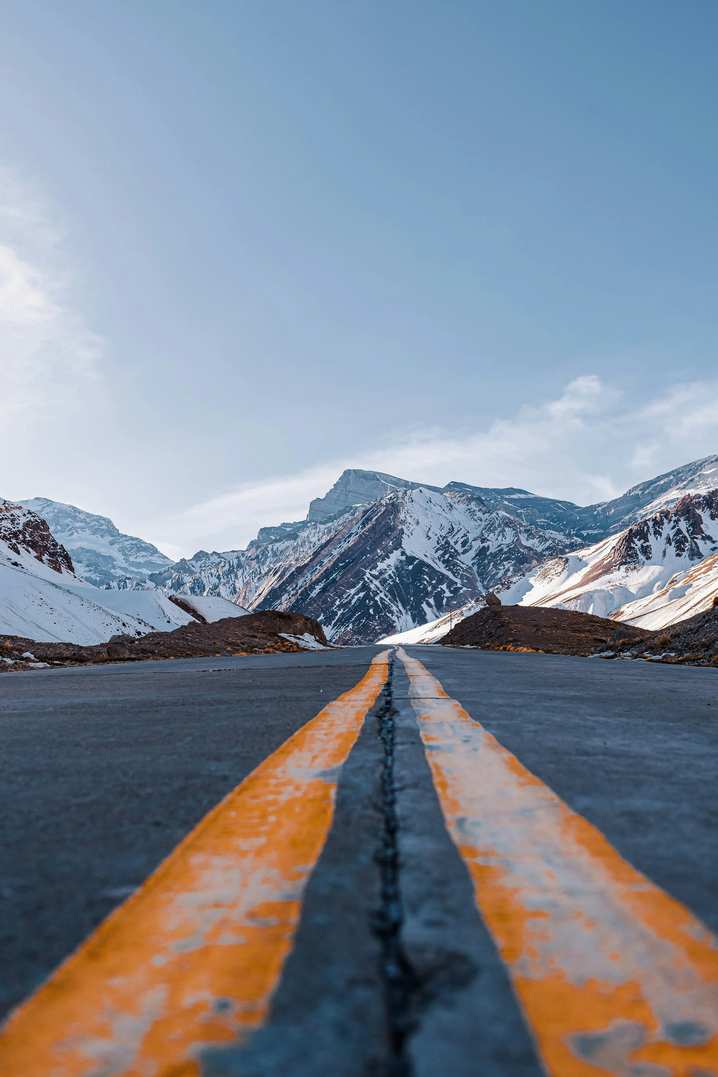 A long straight mountain road with yellow center lines in a snow-covered mountainous landscape, under a blue sky.