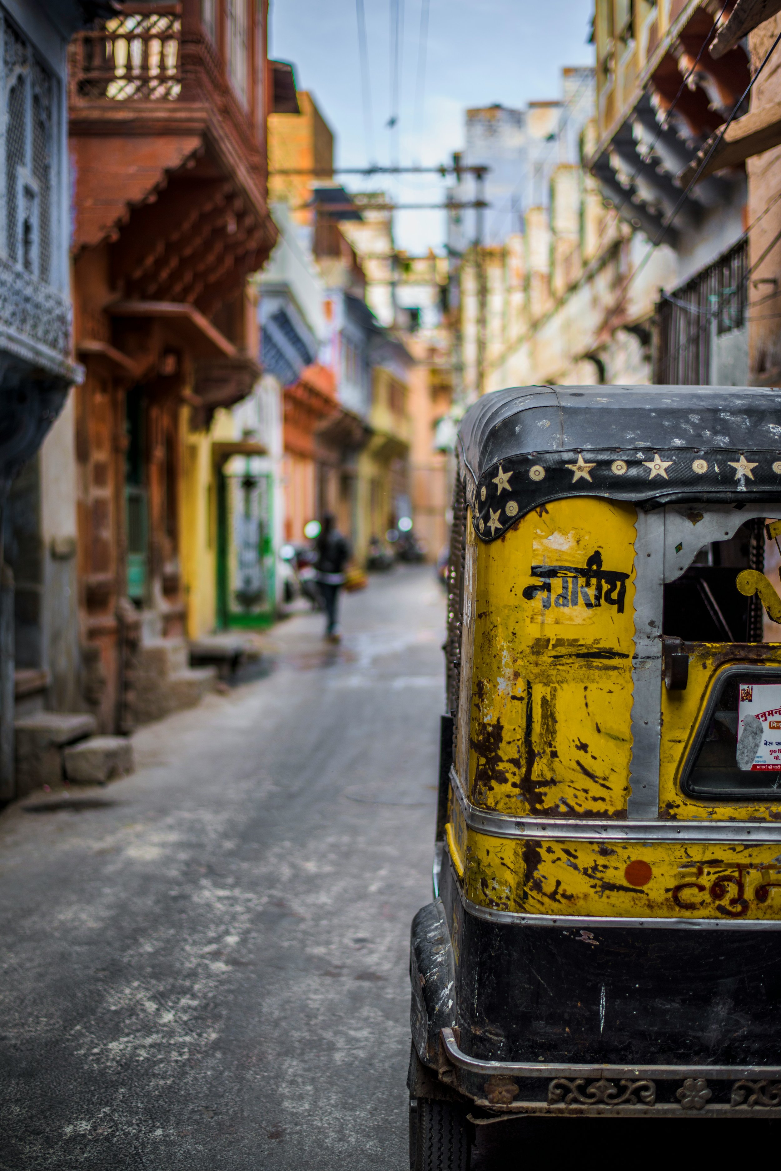 A yellow and black auto rickshaw parked on a street in front of colorful buildings with balconies, wires hanging overhead, and a person walking in the distance.