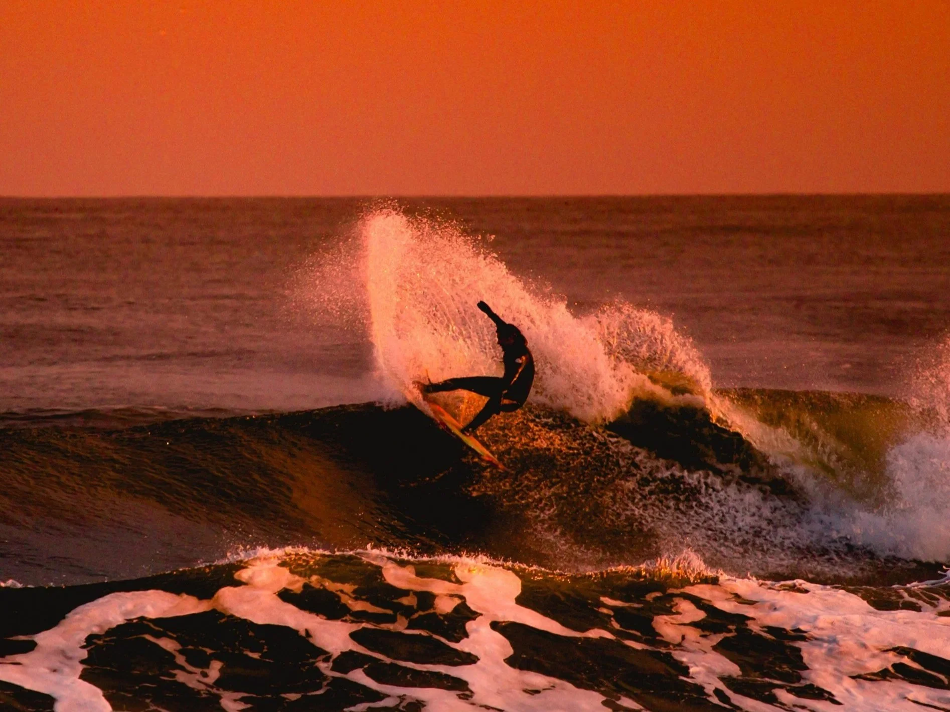 A person surfing on a wave during sunset, with a pink and orange sky, and foamy water in the foreground.