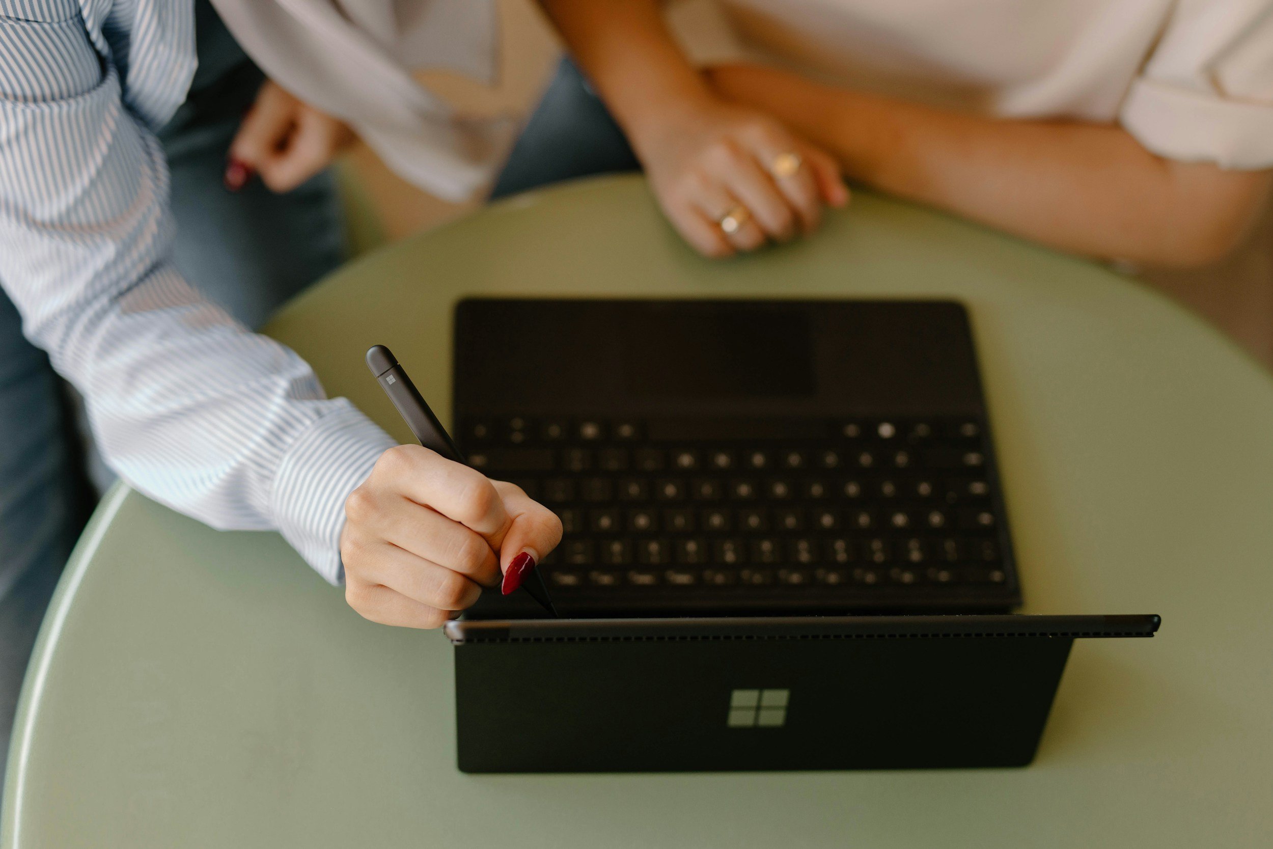 A person with dark red painted nails holding a stylus over a Microsoft Surface tablet. The tablet is on a green table with a black Microsoft Surface laptop in front of it. The person is signing or drawing on the tablet.