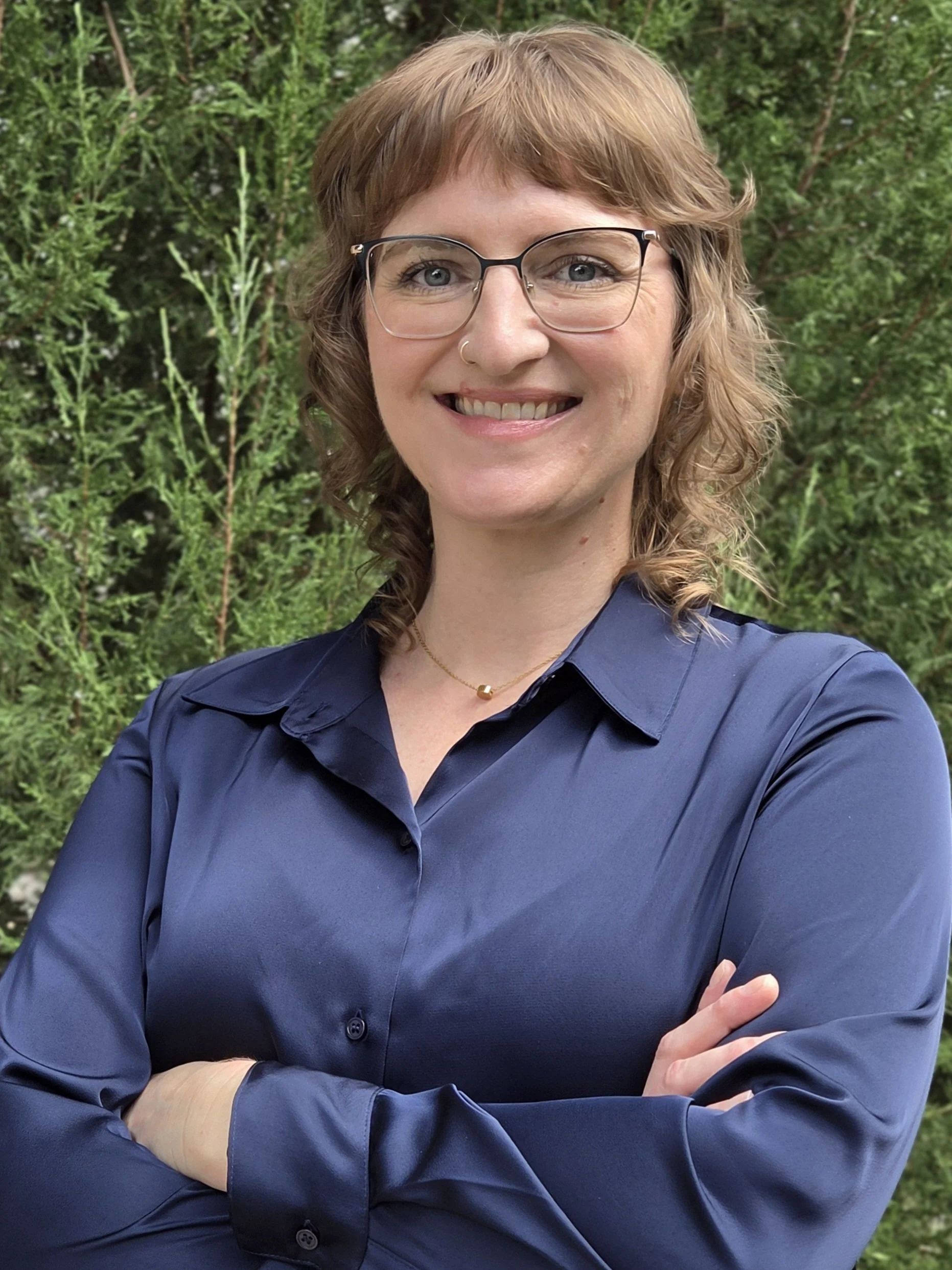A person wearing glasses stands outside in front of a juniper tree. The person is smiling and has a confident stance with their arms folded across their chest.