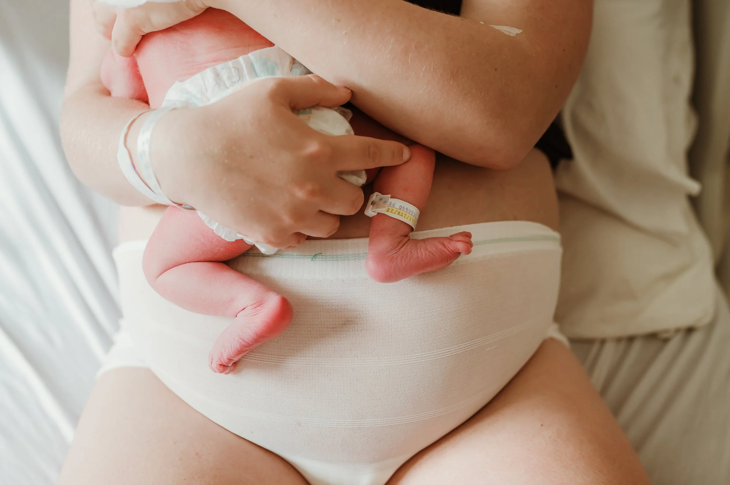 Close-up of a newborn baby lying on a hospital bed, with an adult hand gently holding the baby's tiny foot. The baby is wearing a hospital wristband and diaper, and the scene captures a moment of care and tenderness.