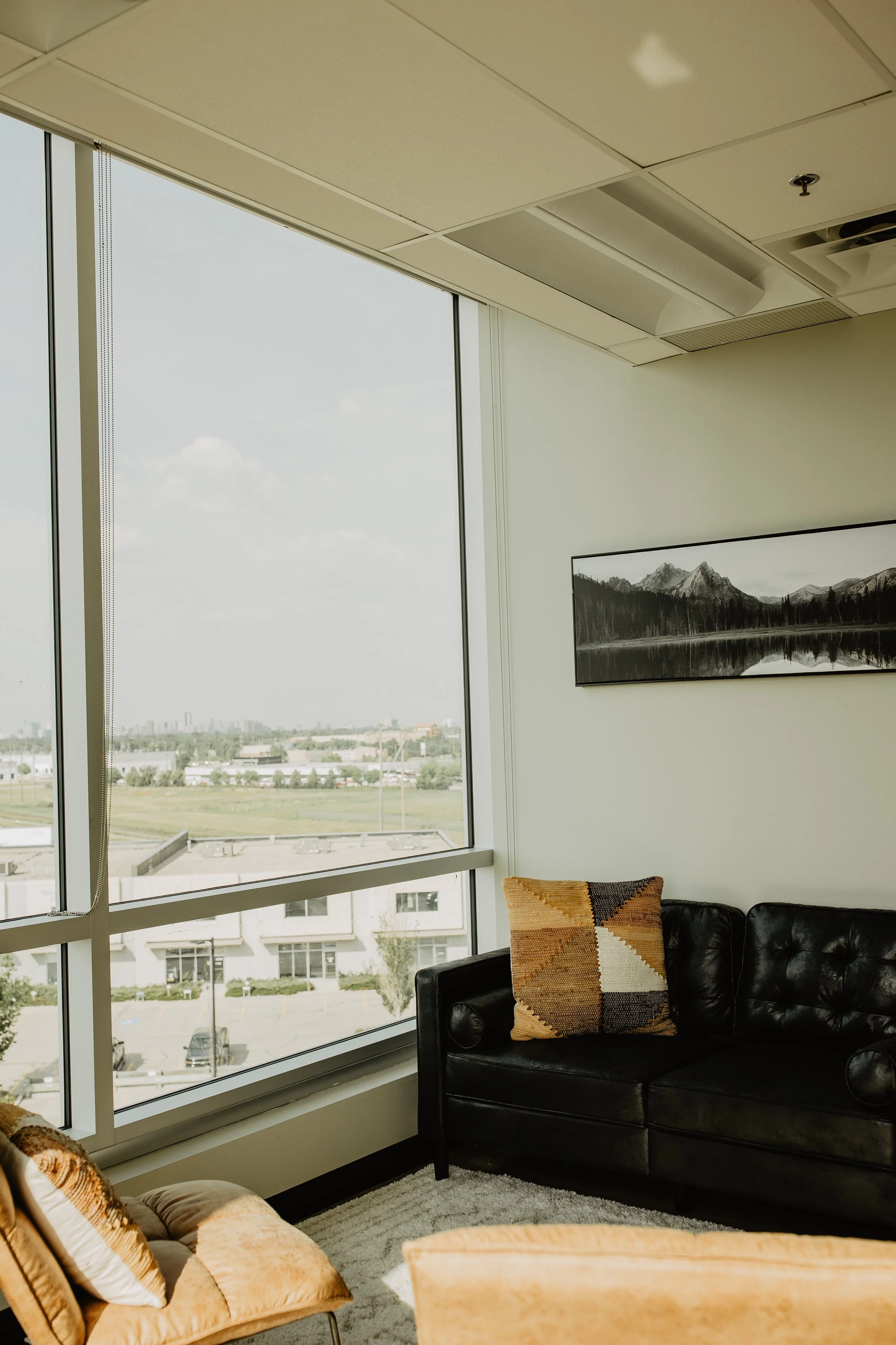 Interior of an office lounge area with large window showing cityscape, black leather sofa with patterned cushion, wall art of mountainous landscape, and beige chair with pillows.