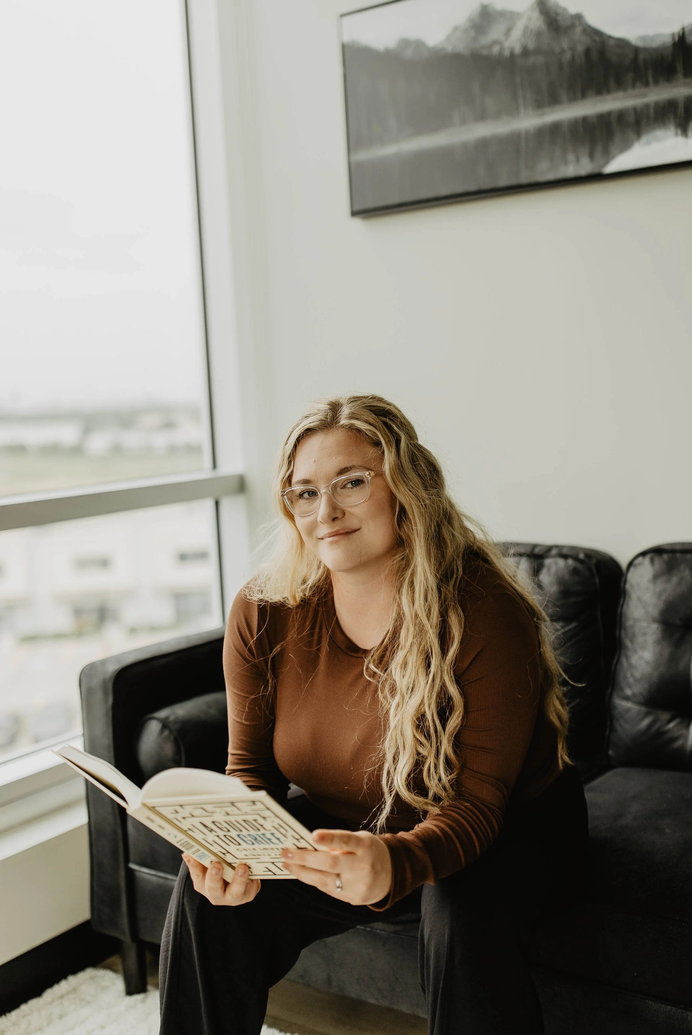 A woman with long wavy blonde hair, glasses, and a brown long-sleeve shirt sitting on a black sofa, reading a book titled 'The Subtle Art of Not Giving a F*ck' in a modern office or lounge area with large window and framed landscape photograph on the wall.