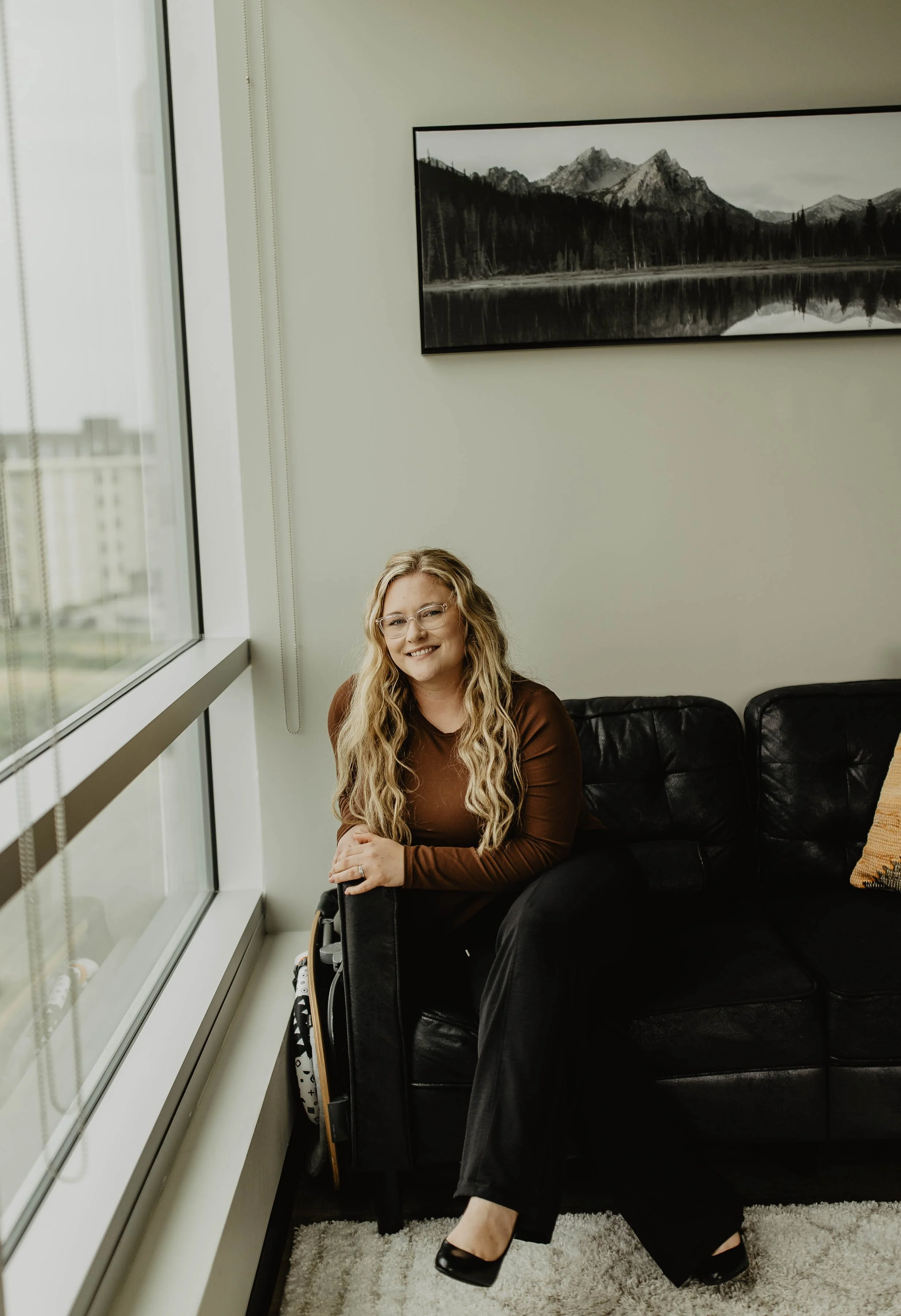 A woman sitting on a black leather couch near a window, smiling, with a mountain landscape painting on the wall behind her. She has long, wavy blonde hair and glasses, wearing a brown top and black pants.