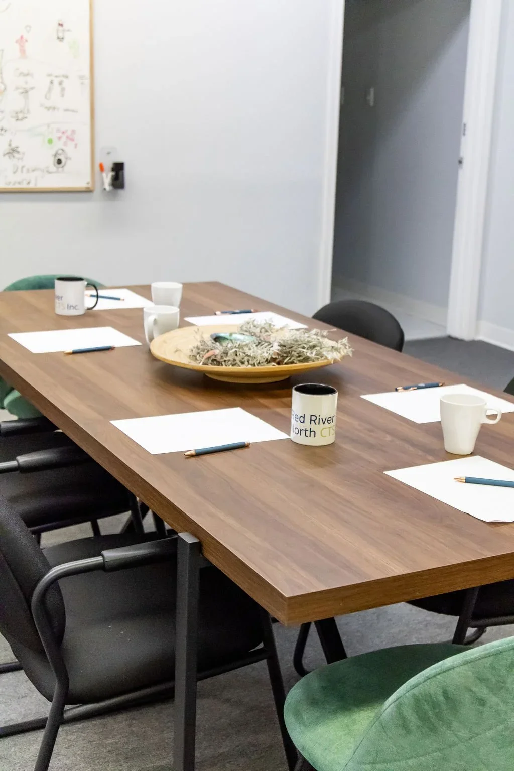 A conference room with a wooden table, six paper pads with pens, and five coffee mugs. A large yellow bowl with dried plant material is in the center of the table. The room has a whiteboard with drawings and writing on the wall, and open doorway leading to another room.