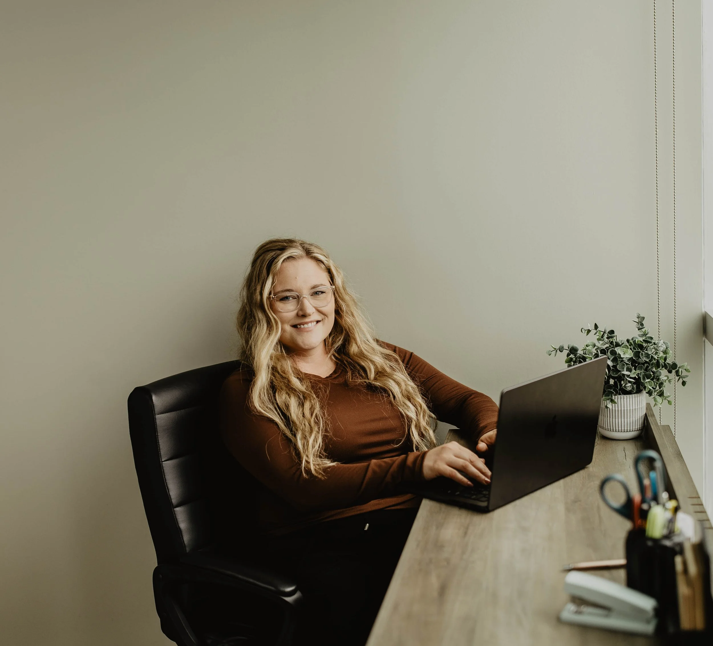 A woman with curly blonde hair and glasses sitting at a desk working on a laptop, with plants and office supplies visible on the desk.