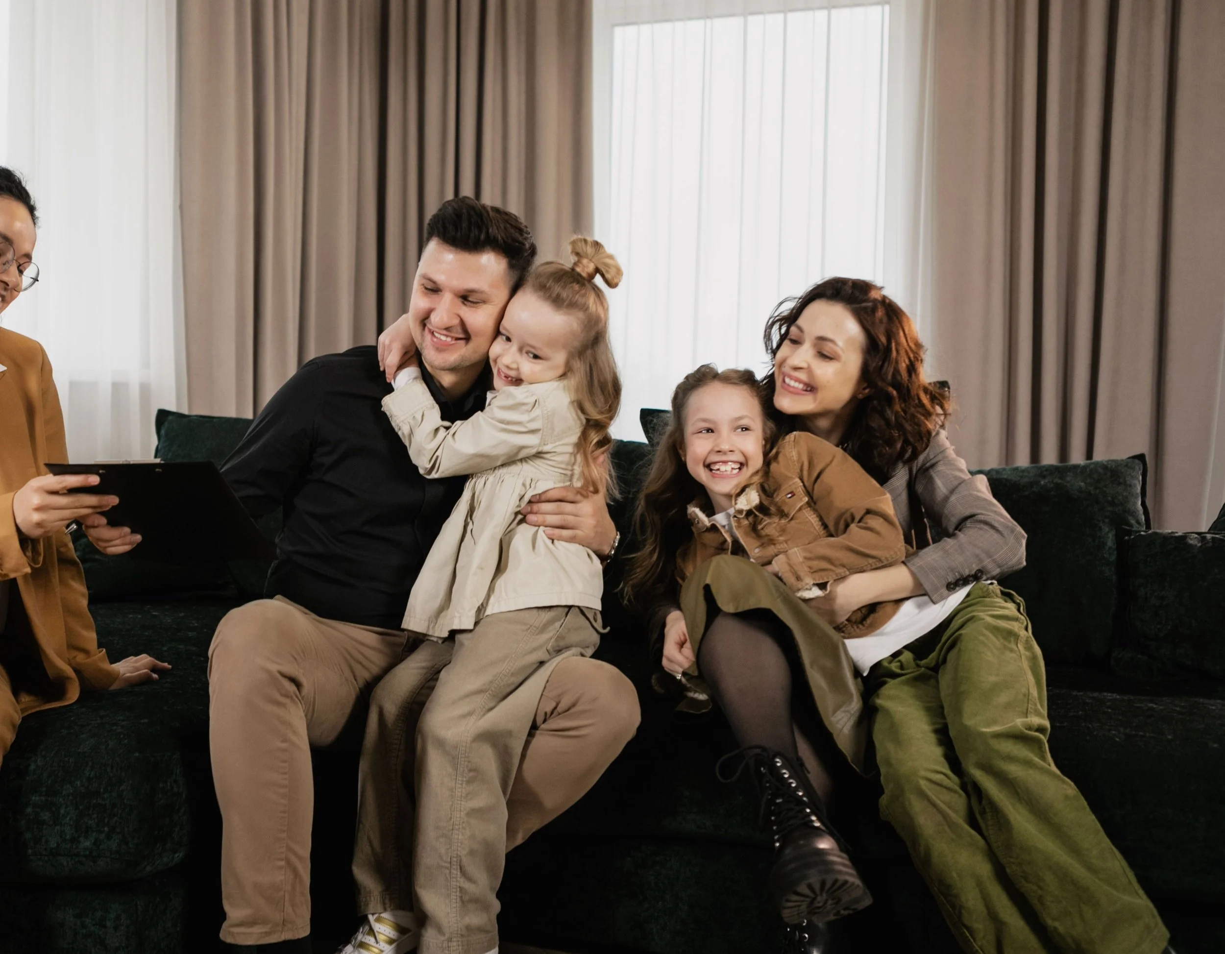 A group of smiling people sitting on a dark carpeted floor in a room with beige curtains and large windows. They are hugging and enjoying each other's company.