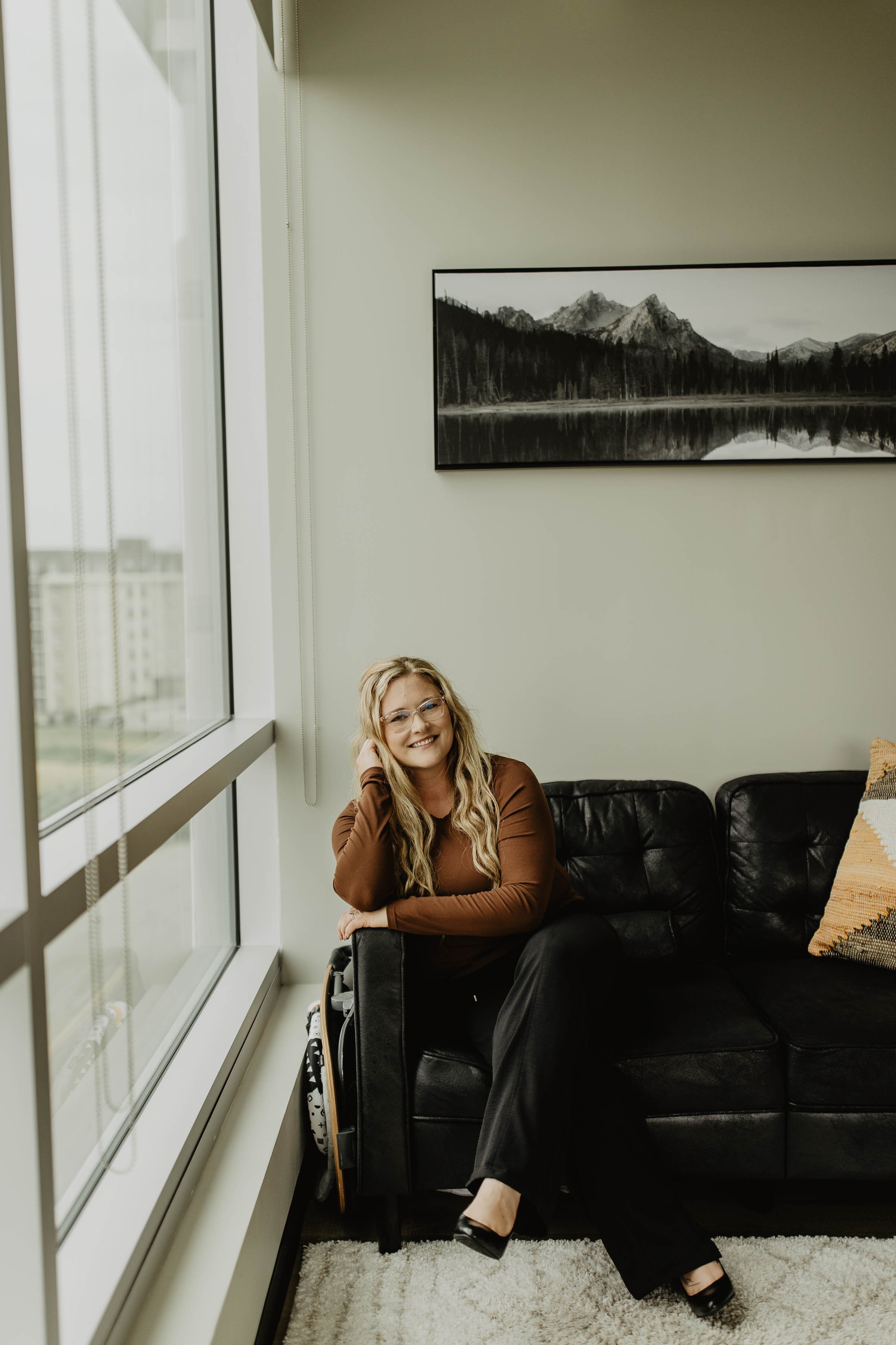 A woman with long curly blonde hair and glasses sitting on a black leather couch near a large window with blinds, smiling at the camera. There is a mountain landscape picture on the wall behind her and a beige pillow with a fringed edge on the couch.