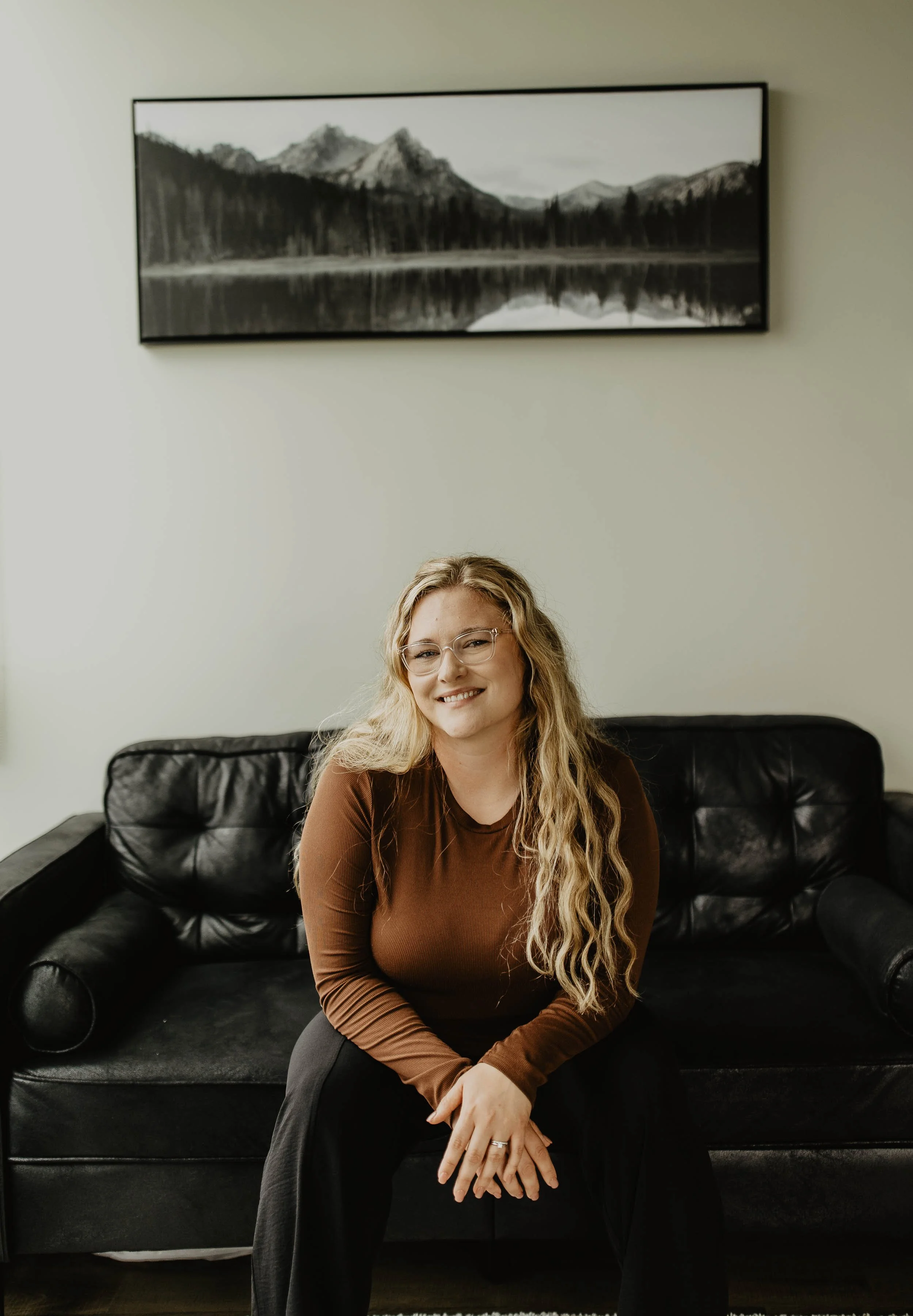 A young woman with long blonde hair and glasses sitting on a black leather sofa, smiling at the camera, in a room with a mountain landscape painting on the wall behind her.