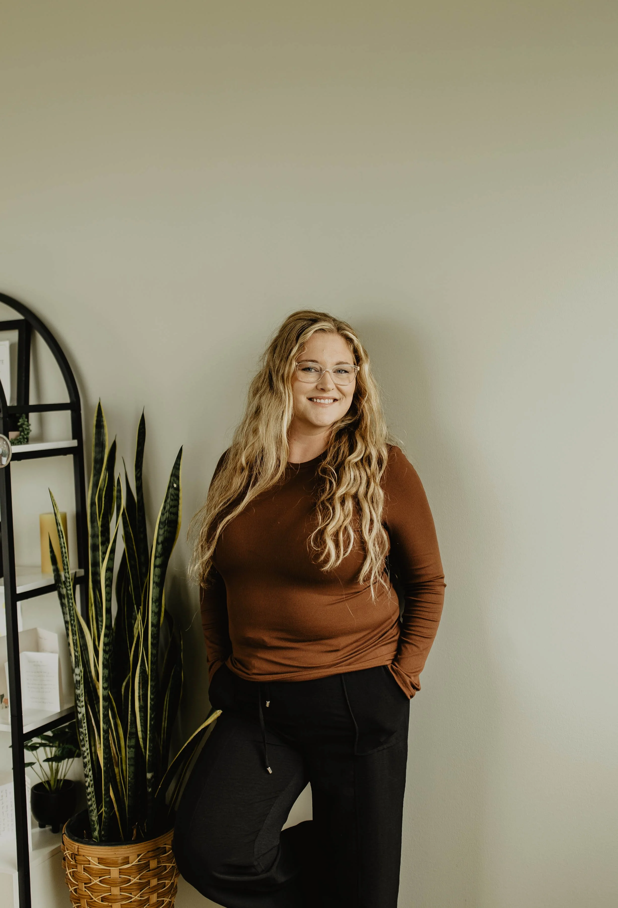 A woman with long blonde hair, glasses, wearing a brown long-sleeve top and black pants, standing indoors next to a white wall and a tall plant in a woven basket.