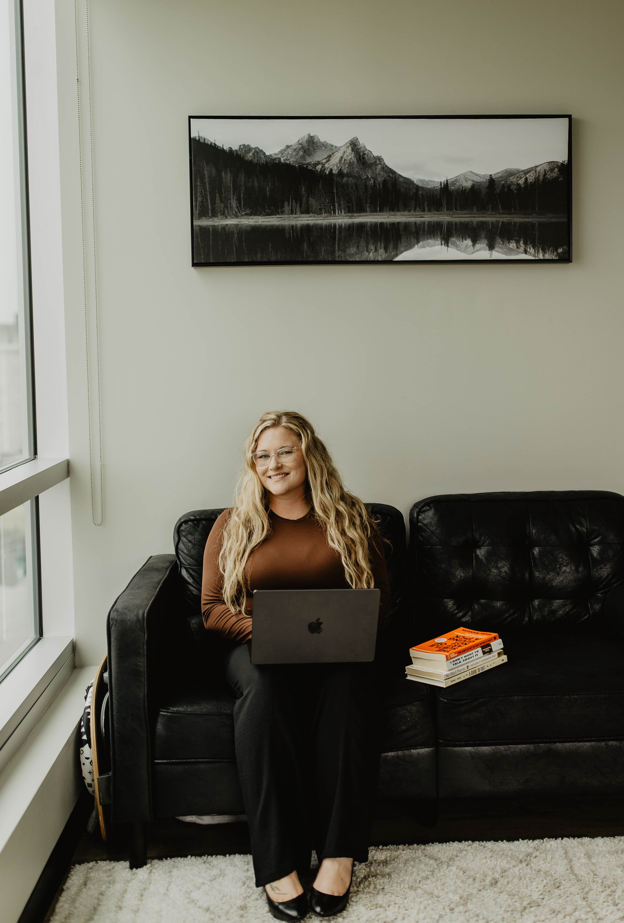 A woman with glasses and long curly blonde hair sitting on a black leather couch, using a laptop. She is smiling and wearing a brown long-sleeve top, black pants, and black flats. There is a stack of books on the coffee table beside her. Behind her, there is a large window on the left and a framed black-and-white mountain landscape photograph on the wall.