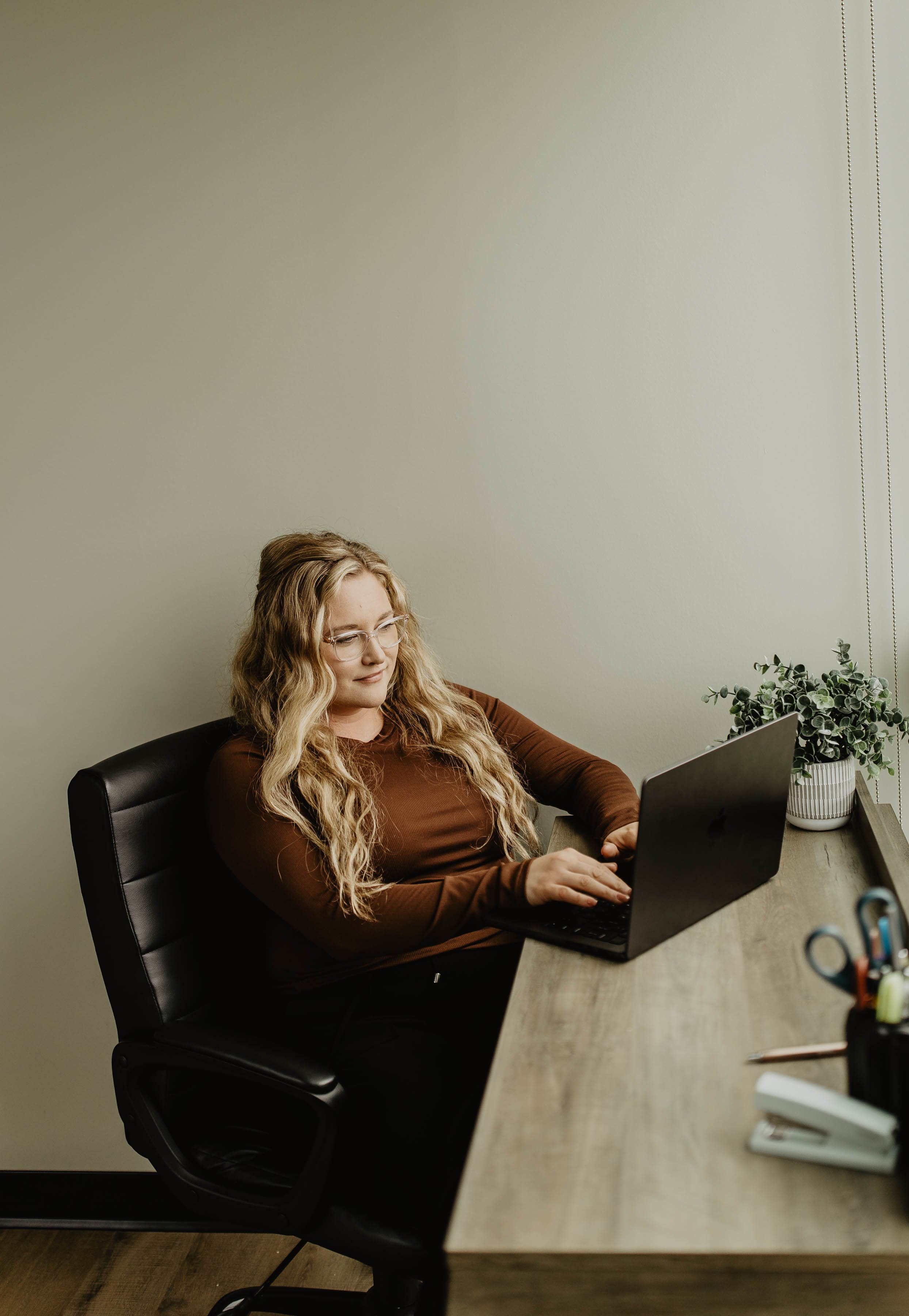 A woman with long blonde hair and glasses sitting at a desk, working on a laptop in an office.