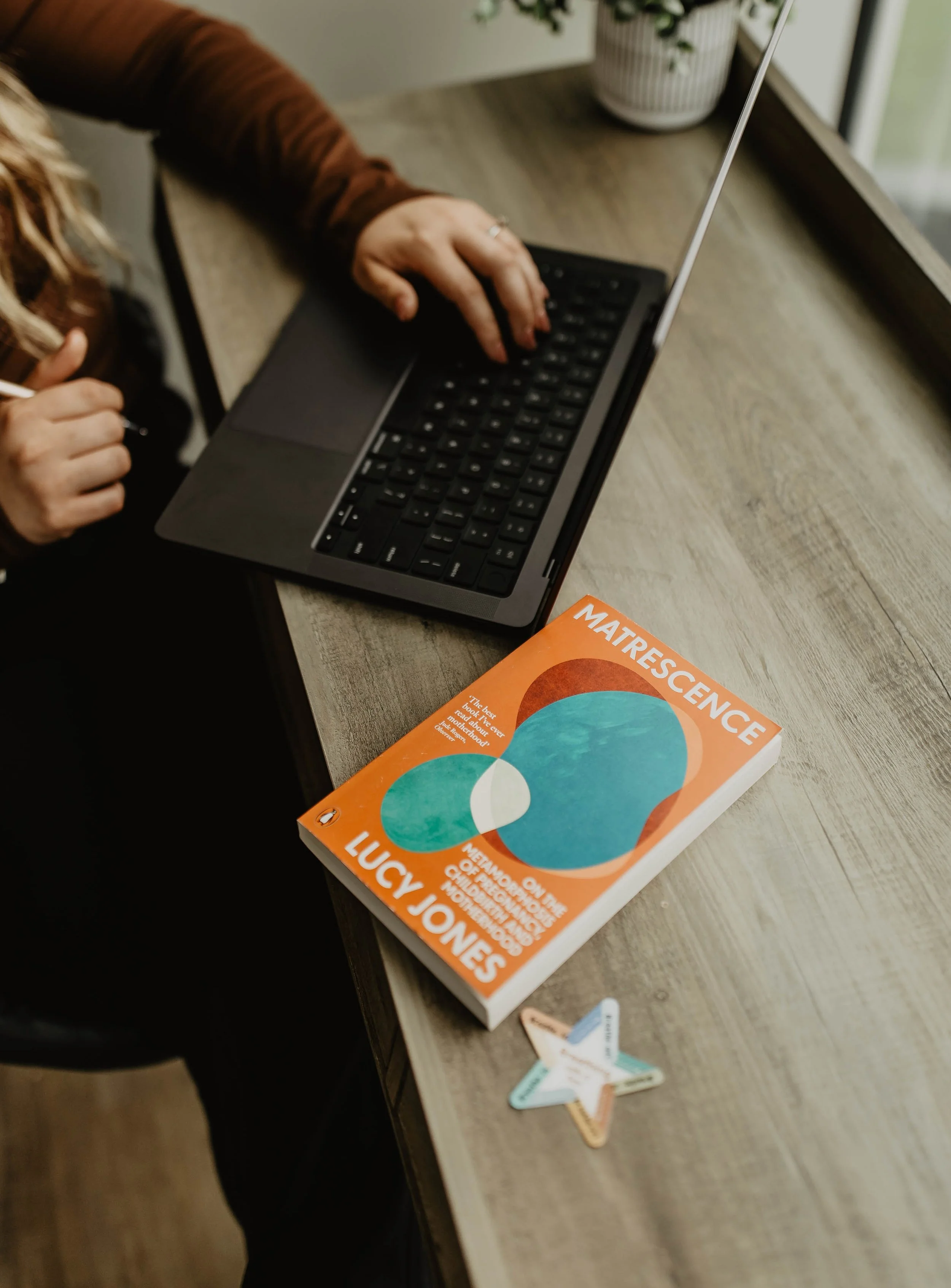 A person working on a laptop at a desk with a book titled "Matrescence" by Lucy Jones and a few star-shaped sticky notes nearby.
