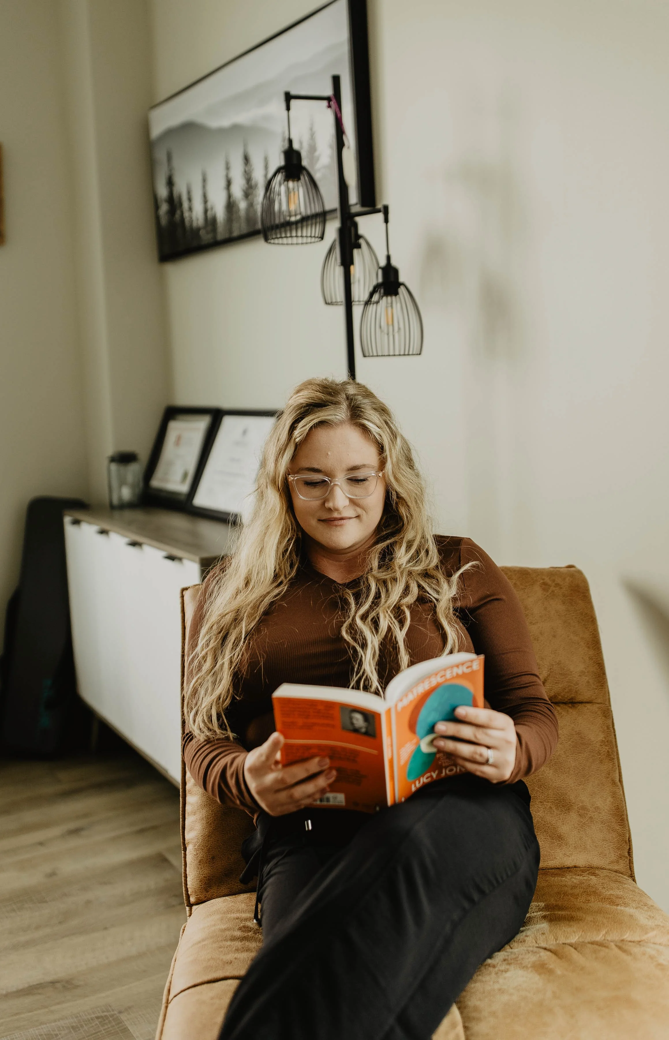 A woman with curly blonde hair and glasses sitting on a brown chair, reading a book titled 'Chemistry' in a cozy living room with wall art, framed photos, and modern lighting fixtures.