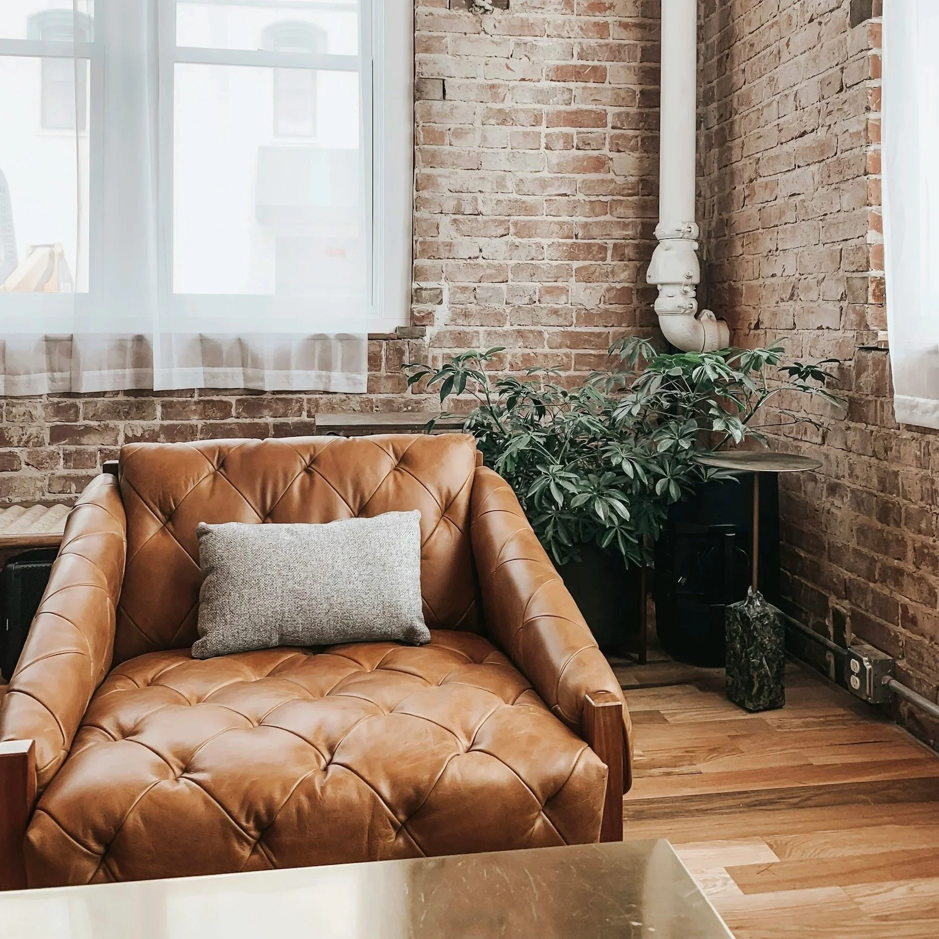 Interior of a room with exposed brick walls, a leather armchair with a cushion, large windows with sheer curtains, a potted plant, and a marble-topped side table.
