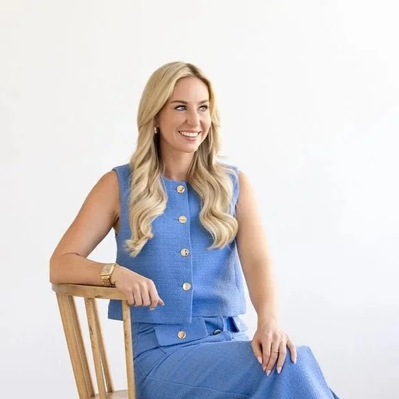 Savannah Goudreau, with long blonde hair wearing a sleeveless blue top and matching skirt, sitting on a wooden chair against a plain white background.