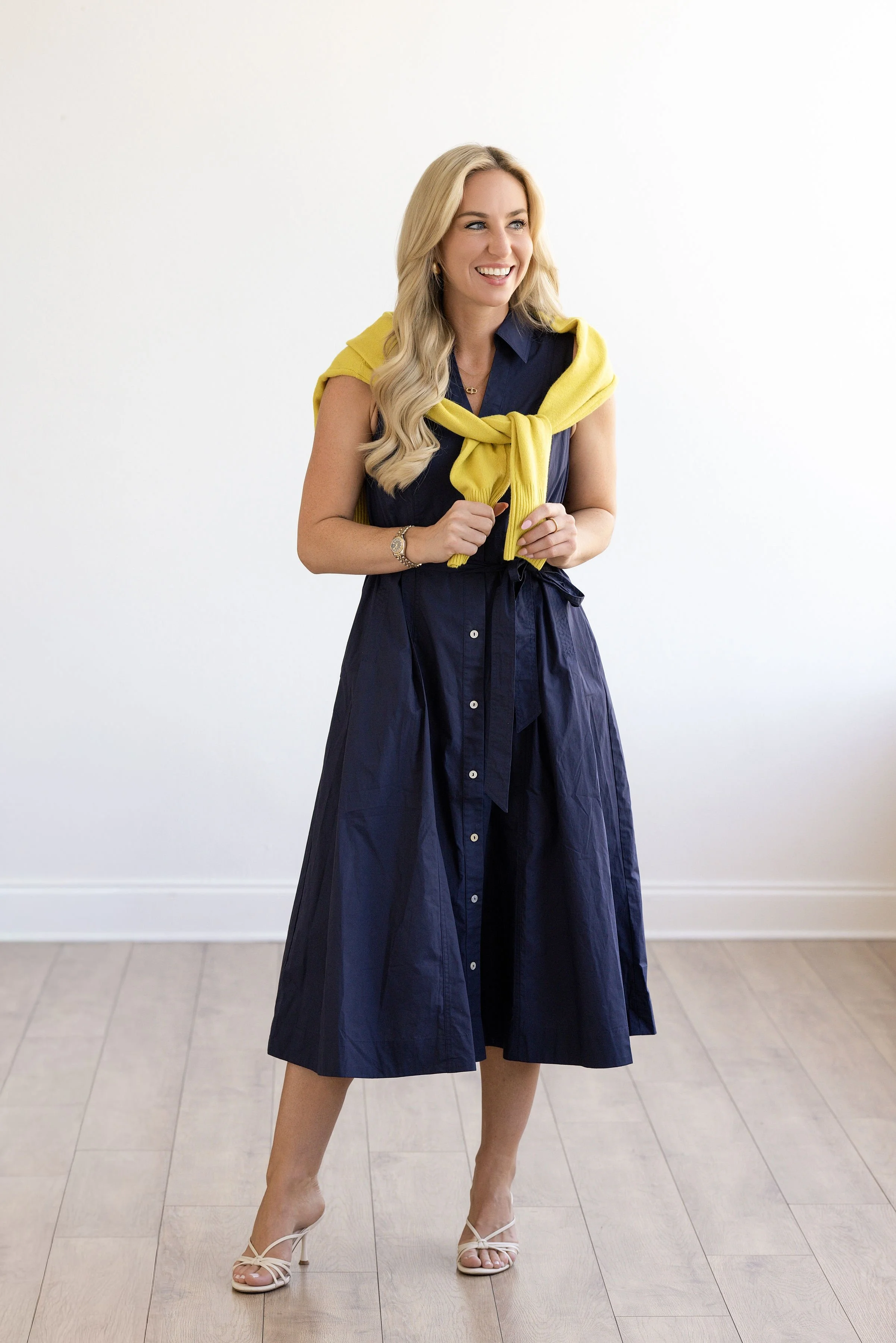 A woman with long blonde hair wearing a navy blue dress with a yellow sweater draped over her shoulders. She is smiling, standing on a light-colored wooden floor against a plain white wall, wearing white high-heeled sandals.