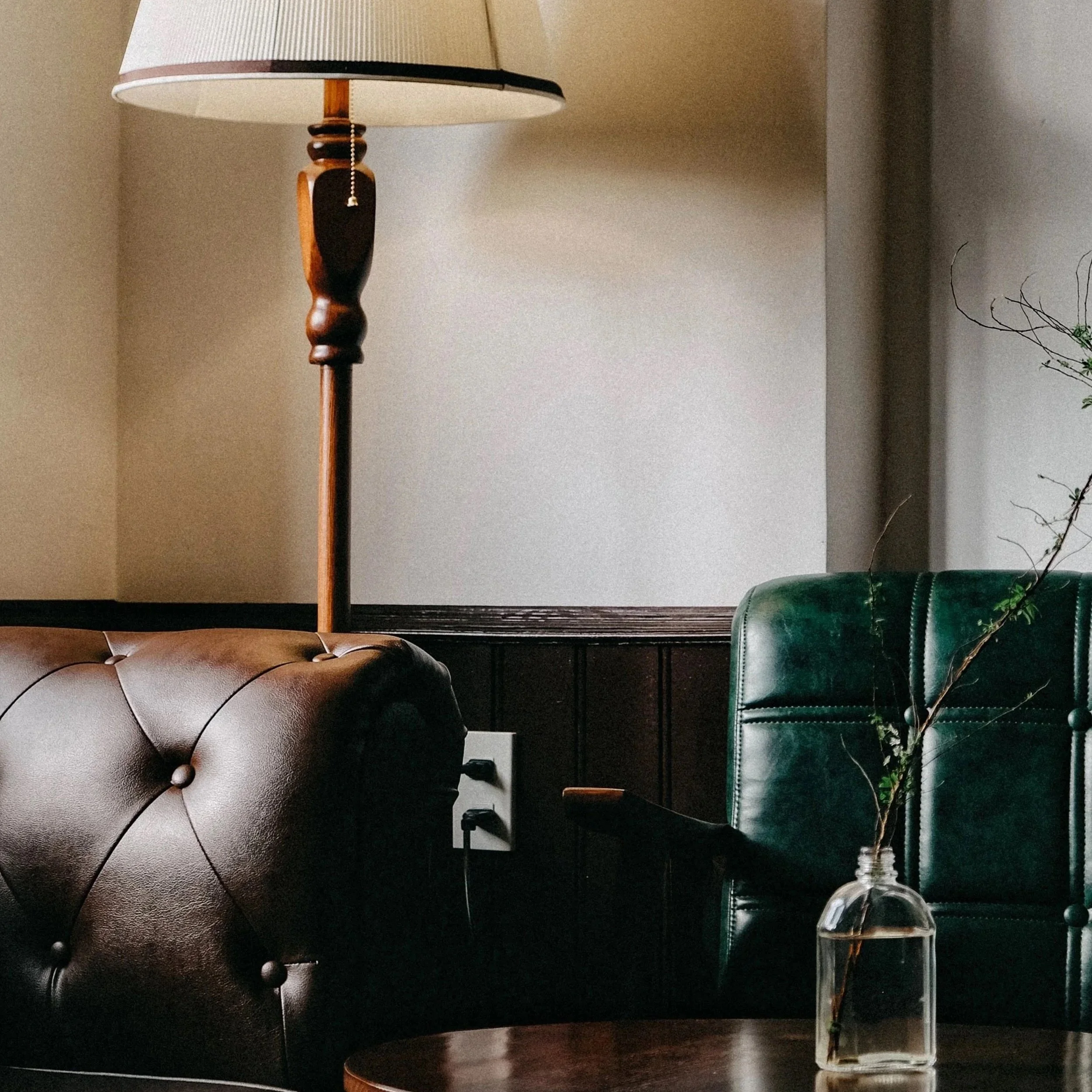 Living room corner with a brown tufted leather armchair, a green leather armchair, a wooden side table, a tall vintage floor lamp with a beige shade, and a glass vase with twisted twigs.